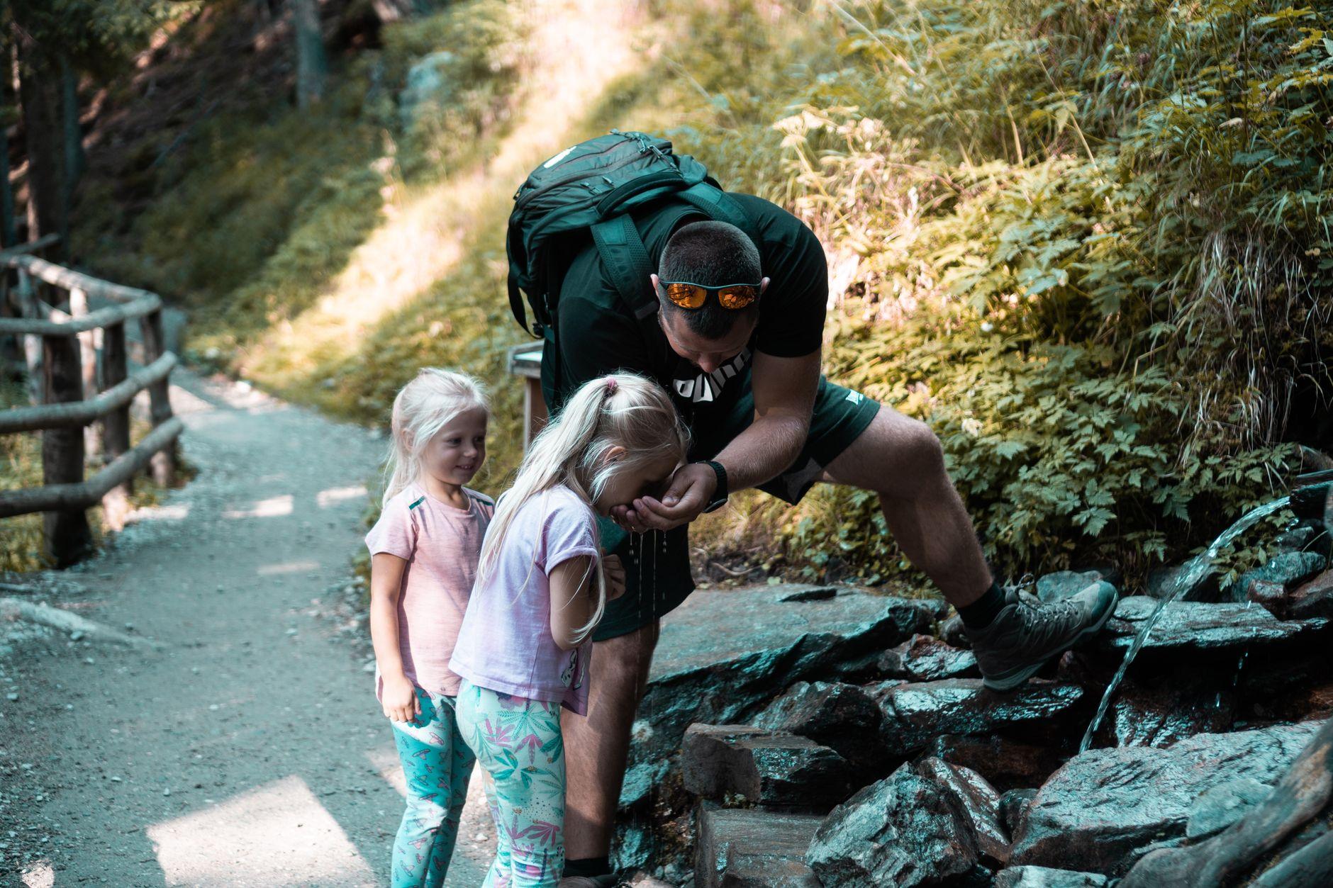 A father helping his young daughter drink from a natural spring along a forest trail